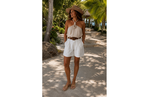 Woman in a white shorts taupe Satin top wearing a straw hat standing on a tropical path with palm trees.